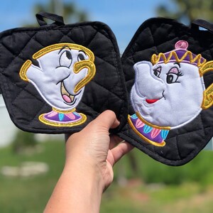 K&ouml;nnte beinhalten: Ein Paar schwarze, gesteppte Topflappen mit gestickten Bildern von Chip und Mrs. Potts aus Disneys Die Sch&ouml;ne und das Biest. Chip ist eine Teetasse mit gelbem Rand und Mrs. Potts ist eine Teekanne mit einer goldenen Krone.