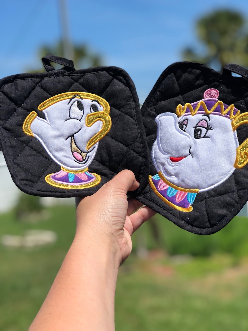 K&ouml;nnte beinhalten: Ein Paar schwarze, gesteppte Topflappen mit gestickten Bildern von Chip und Mrs. Potts aus Disneys Die Sch&ouml;ne und das Biest. Chip ist eine Teetasse mit gelbem Rand und Mrs. Potts ist eine Teekanne mit einer goldenen Krone.