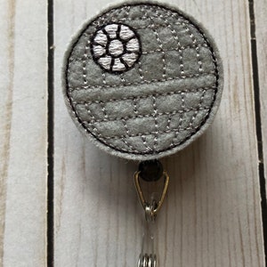 Könnte beinhalten: Ein grauer Filz-Badgehalter in Form des Todessterns mit einem gestickten Design in Silber und Schwarz. Der Badgehalter ist an einer durchsichtigen, einziehbaren Badge-Spule mit einem silbernen Metallclip befestigt.