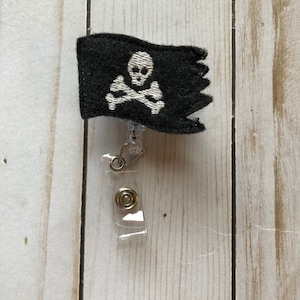 Könnte beinhalten: Eine schwarze Stoff-Piratenflagge mit einem weißen Totenkopf- und Knochenkreuz-Design, das an einem durchsichtigen, einziehbaren Badge-Halter befestigt ist.