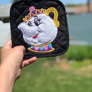 K&ouml;nnte beinhalten: Schwarzer gesteppter Topflappen mit einem gestickten Bild von Mrs. Potts, der Teekannenfigur aus Die Sch&ouml;ne und das Biest. Mrs. Potts tr&auml;gt eine lila und goldene Krone und hat ein wei&szlig;es Gesicht mit rosa Wangen und roten Lippen.