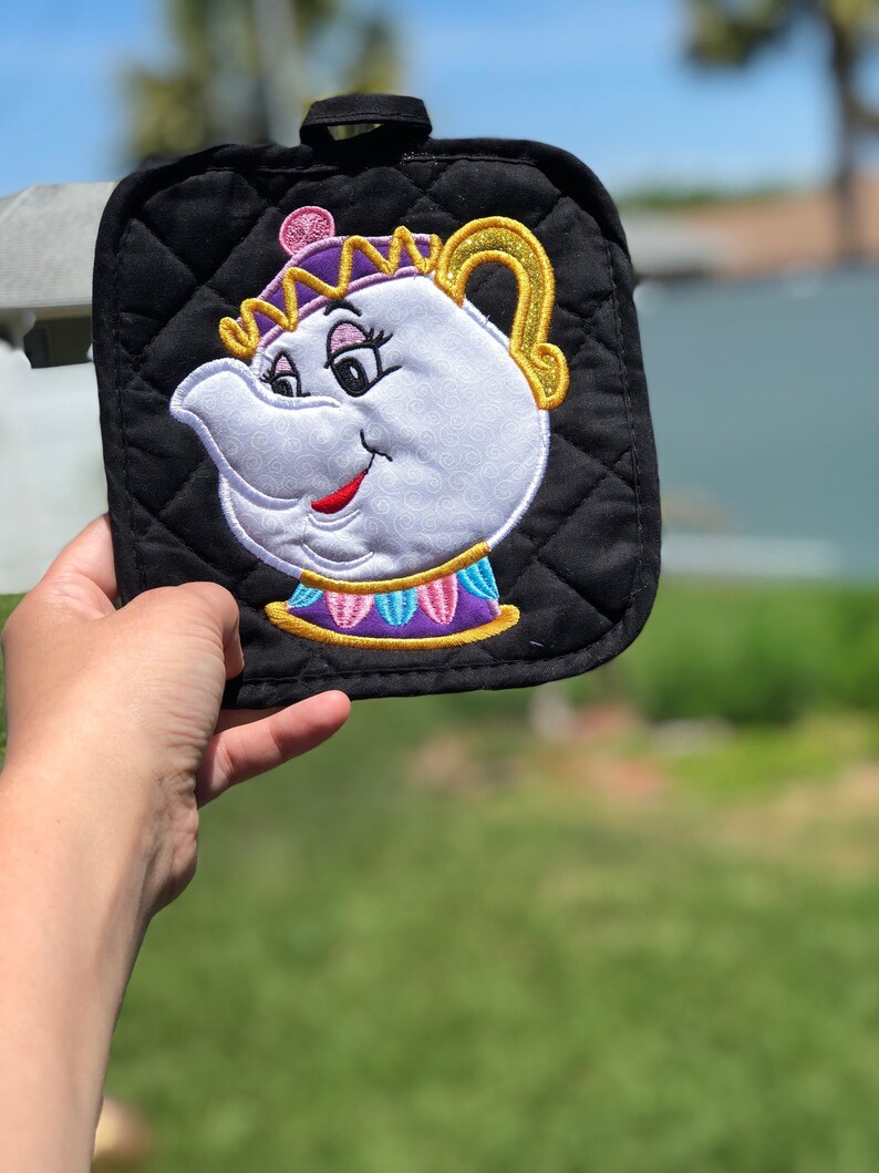 K&ouml;nnte beinhalten: Schwarzer gesteppter Topflappen mit einem gestickten Bild von Mrs. Potts, der Teekannenfigur aus Die Sch&ouml;ne und das Biest. Mrs. Potts tr&auml;gt eine lila und goldene Krone und hat ein wei&szlig;es Gesicht mit rosa Wangen und roten Lippen.