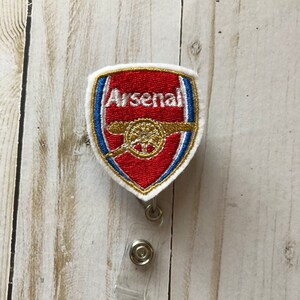 Könnte beinhalten: Weißer Stoff-Badgehalter mit einem gestickten Arsenal Football Club Logo in Rot, Weiß, Gold und Blau. Der Badgehalter hat einen transparenten, einziehbaren Badgeclip aus Kunststoff.