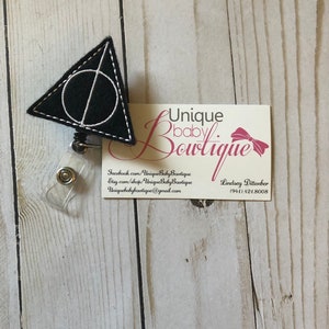 Könnte beinhalten: Schwarz-weiß gesticktes Abzeichen mit einem Dreieck- und Kreisdesign. Das Abzeichen ist an einem durchsichtigen, einziehbaren Plastik-Ausweishalter befestigt. Das Abzeichen ist mit "Unique baby Boutique" sowie einer Website und einer Telefonnummer beschriftet.