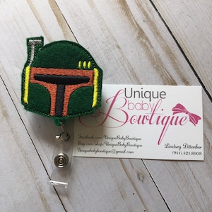 Könnte beinhalten: Ein gesticktes Abzeichen mit dem Helm eines Star Wars-Charakters in Grün, Orange und Gelb. Das Abzeichen ist an einem durchsichtigen, einziehbaren Ausweishalter aus Kunststoff befestigt. Der Ausweishalter ist an einer Visitenkarte mit dem Text "Unique baby Boutique" und Kontaktinformationen befestigt.