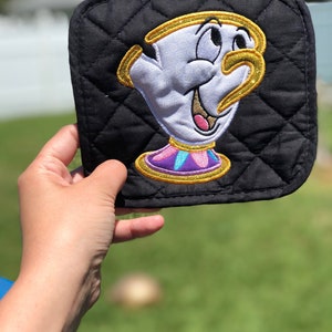 K&ouml;nnte beinhalten: Schwarzer gesteppter Topflappen mit einem goldenen gestickten Bild von Chip aus Die Sch&ouml;ne und das Biest. Chip l&auml;chelt und hat einen goldenen Griff.