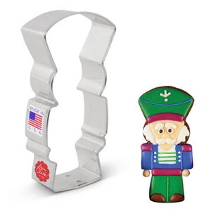 May include: A stainless steel cookie cutter in the shape of a nutcracker soldier, next to a decorated cookie of the same design. The cookie is green, blue, red, and brown. The cookie cutter has a label that says "Made in USA".