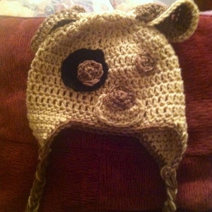 May include: A crocheted hat with brown and beige yarn, featuring a large brown eye and smaller brown accents. The hat has long ties that can be tied under the chin.
