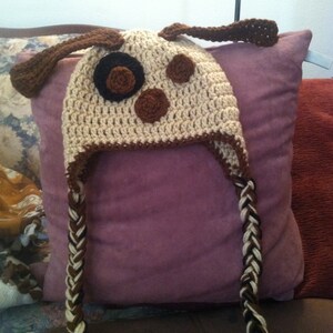 May include: A crocheted dog hat with brown and white ear flaps and a brown and white braided strap. The hat has brown felt spots on the crown.