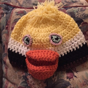 May include: A crocheted yellow duck hat with black, white, and brown stripes. The hat has a large orange beak and two eyes with black and pink details.