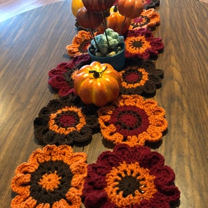 May include: A handmade crochet table runner featuring a floral design in shades of orange, brown, and burgundy. The runner is decorated with artificial pumpkins and gourds, creating a fall-themed centerpiece for a dining table.