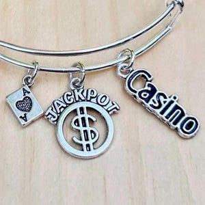 May include: Silver charm bracelet with four charms: a playing card with a heart, a dollar sign with the word "JACKPOT" around it, a silver charm with the word "Casino" on it, and a silver charm with a heart.