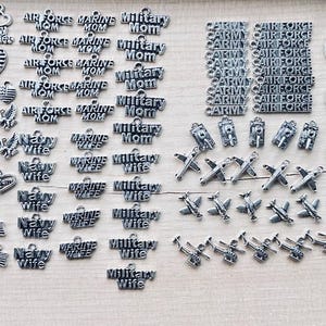 May include: A collection of silver-toned charms and pendants. Designs include hearts with stripes, military emblems, and text such as "Air Force Mom" and "Military Wife". Various shapes and sizes are present.