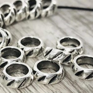 May include: Close-up of several silver-colored metal beads with a textured, diagonal pattern. The beads are round with a hole in the center, and are likely used for jewelry making or crafting. The beads are scattered on a light-colored wooden surface.