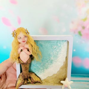 May include: A small sculpture of a mermaid with long blonde hair and a metallic gold tail, positioned in front of a square white frame containing a textured depiction of a sandy beach with blue ocean waves and a small white starfish in the corner.