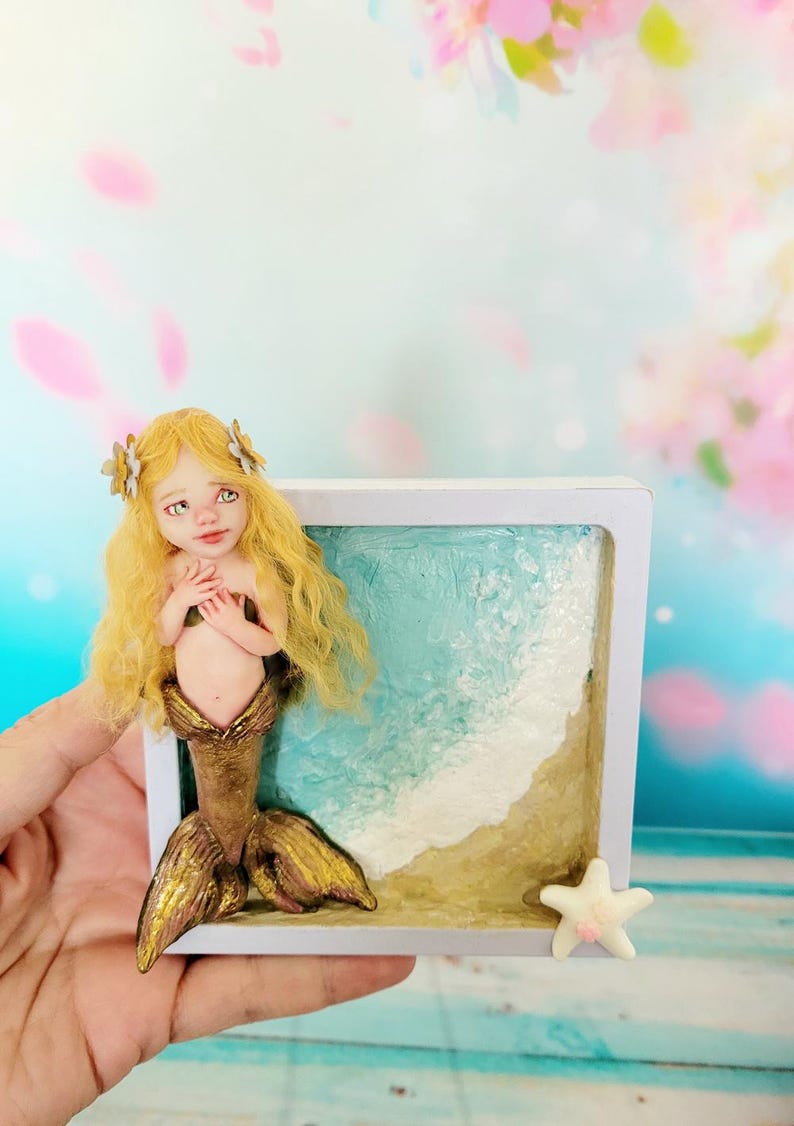 May include: A small sculpture of a mermaid with long blonde hair and a metallic gold tail, positioned in front of a square white frame containing a textured depiction of a sandy beach with blue ocean waves and a small white starfish in the corner.