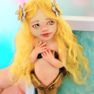 May include: A mermaid figurine with long wavy yellow hair adorned with two small white flowers. The figure has pale skin, green eyes, and a metallic gold tail. The hands are positioned over the chest, and the facial features are painted with detail.