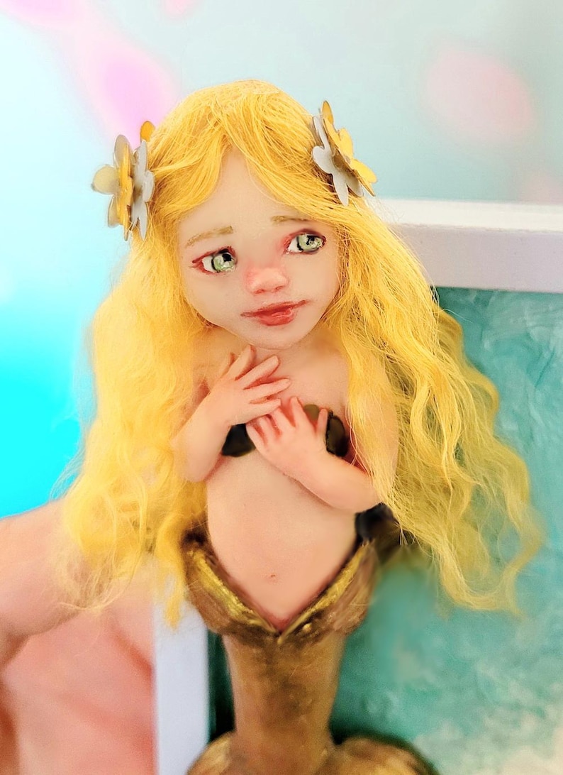 May include: A mermaid figurine with long wavy yellow hair adorned with two small white flowers. The figure has pale skin, green eyes, and a metallic gold tail. The hands are positioned over the chest, and the facial features are painted with detail.