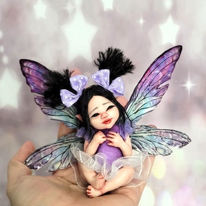 Könnte beinhalten: Eine kleine, handgefertigte Feenpuppe mit lila Flügeln, schwarzen Haaren und einem lila Kleid. Die Fee sitzt in einer Hand vor einem funkelnden Hintergrund.