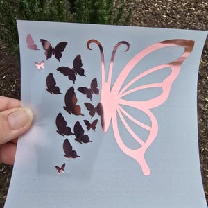 May include: A decorative butterfly design in rose gold and brown. The design features a large butterfly wing on the right and a cluster of smaller butterflies on the left, all on a light gray background.