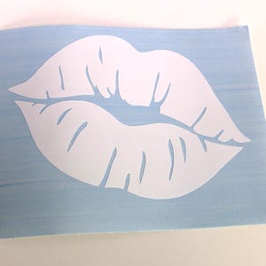 May include: A white lip-shaped decal on a light blue background. The decal is a cut-out of a pair of lips, with details showing the lip lines. The background is a light blue rectangle.