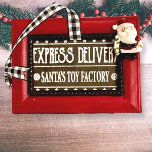 May include: A red rectangular Christmas ornament with the text "EXPRESS DELIVERY" and "SANTA'S TOY FACTORY". A small Santa Claus figure is on the right side. The ornament has a black and white checkered ribbon for hanging.