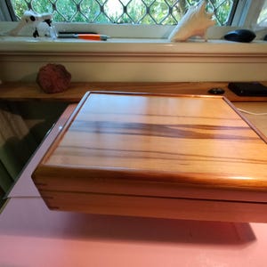 May include: A rectangular wooden box with a striped pattern on the lid. The box is made of light and dark brown wood and has rounded corners. The box is sitting on a pink surface.