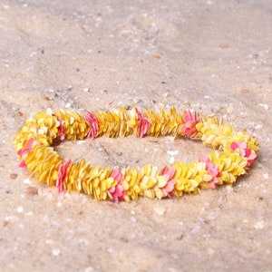 Puede incluir: Un collar de flores amarillas y rosas descansa sobre arena. El collar est&aacute; compuesto por numerosos elementos peque&ntilde;os en forma de p&eacute;talo, creando una forma circular. La arena es de color marr&oacute;n claro y ligeramente texturizada.