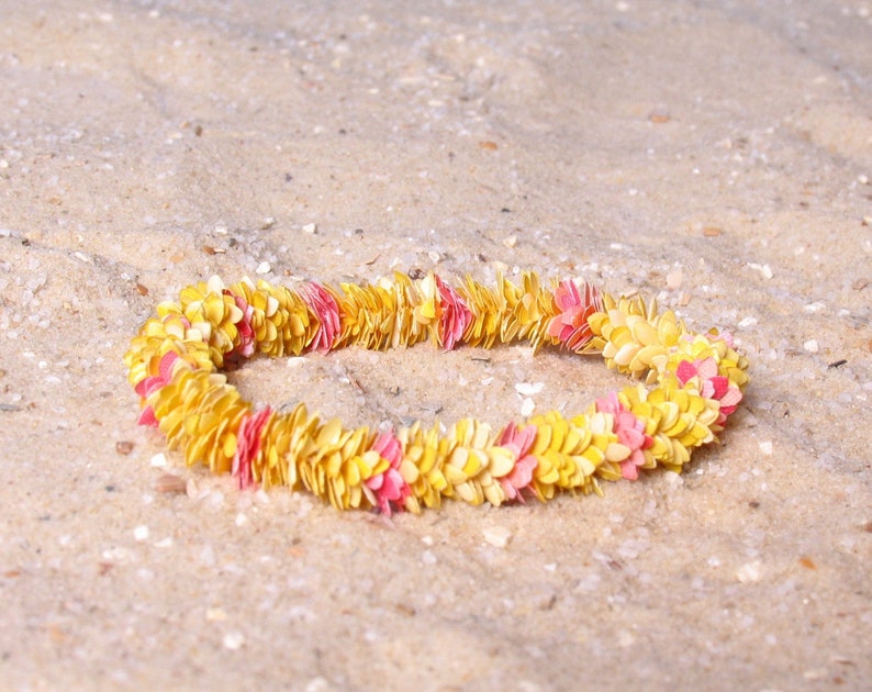 Puede incluir: Un collar de flores amarillas y rosas descansa sobre arena. El collar est&aacute; compuesto por numerosos elementos peque&ntilde;os en forma de p&eacute;talo, creando una forma circular. La arena es de color marr&oacute;n claro y ligeramente texturizada.