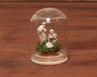 May include: A miniature nativity scene enclosed in a clear glass dome with a light wood base. Inside, two off-white figurines stand over a small white bundle resting on a bed of green moss. The dome is rounded at the top and sits on a flat base.
