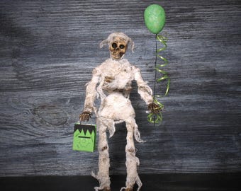 May include: A standing skeleton figure wrapped in tattered off-white gauze bandages. The figure holds a bright green balloon with a curly green ribbon in one hand and a small green gift bag featuring a black-haired monster face in the other.