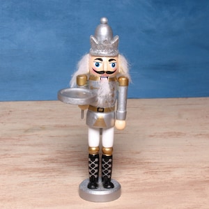 Könnte beinhalten: Eine Nussknacker-Figur mit silberner Krone, silberner Jacke mit goldenen Akzenten und weißer Hose. Die Figur hat weißes Haar und einen Bart, schwarze Stiefel mit weißer Schnürung und hält ein kleines silbernes, rundes Tablett in der rechten Hand.