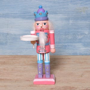 Könnte beinhalten: Eine hölzerne Nussknacker-Figur mit einer hellblauen Krone mit violetten Akzenten, einer rosa Jacke mit hellblauer Verzierung und gestreiften Hosen. Die Figur hat einen weißen Kunstfellbart, klare Strasssteine und hält ein kleines weißes Tablett.