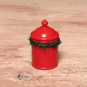 Könnte beinhalten: Ein kleiner, roter Topf mit Deckel und einem grünen Girlande um den Sockel. Der Topf hat eine abgerundete Oberseite und einen kleinen Knopf. Der Hintergrund ist eine hellbraune Holzoberfläche.