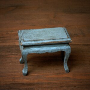 Op de afbeelding: Een kleine tafel in antieke stijl met een rechthoekig blad en gebogen poten. De tafel is geschilderd in een verweerde blauwgrijze kleur, met zichtbare houtnerf. De tafel is ongeveer 15 cm lang, 10 cm breed en 10 cm hoog.
