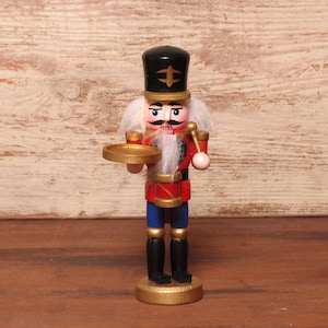 Könnte beinhalten: Eine hölzerne Nussknacker-Figur mit schwarzem Hut und goldenem Emblem, roter Jacke mit Goldverzierungen, blauer Hose und schwarzen Stiefeln. Die Figur hat weißes Haar und einen Bart und hält ein kleines goldenes Tablett in der Hand.