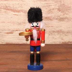 Puede incluir: Una figura de cascanueces de madera con un sombrero negro alto y peludo, chaqueta roja con detalles dorados, pantalones azules y botas negras. La figura tiene barba blanca, bigote pintado y sostiene una pequeña bandeja circular dorada.