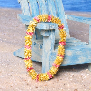 Puede incluir: Un colorido lei hecho de p&eacute;talos amarillos y rosas descansa sobre una silla de madera azul claro en una playa arenosa. El lei es una guirnalda hawaiana tradicional, a menudo usada para celebraciones.