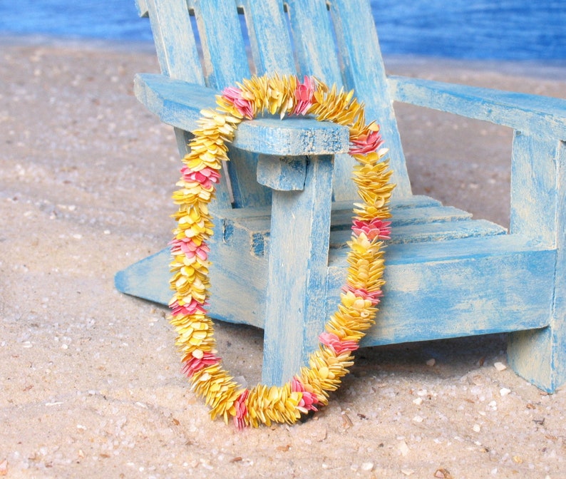 Puede incluir: Un colorido lei hecho de p&eacute;talos amarillos y rosas descansa sobre una silla de madera azul claro en una playa arenosa. El lei es una guirnalda hawaiana tradicional, a menudo usada para celebraciones.