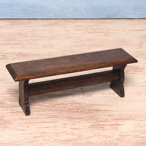 Könnte beinhalten: Eine Miniatur-Holzbank mit dunkelbraunem Finish. Die Bank hat eine rechteckige Sitzfläche und einen unteren Stützbalken. Die Beine sind mit dekorativen Ausschnitten geformt. Diese kleine Bank eignet sich für Puppenhäuser oder andere Miniaturumgebungen.