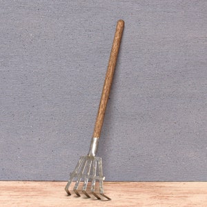 Könnte beinhalten: Ein kleiner Rechen mit einem Holzstiel und Metallzinken. Der Rechen ist aus Metall und Holz und hat einen braunen Holzstiel.