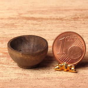 Può includere: Una piccola ciotola di legno marrone scuro si trova su una superficie di legno chiaro accanto a una moneta da 1 centesimo di euro. La moneta è su un supporto dorato.