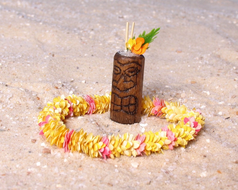 Puede incluir: Un vaso de c&oacute;ctel de madera tallada al estilo tiki, adornado con una flor y vegetaci&oacute;n, y dos pajitas. Est&aacute; rodeado por un lei de flores amarillas y rosas, todo sobre arena.