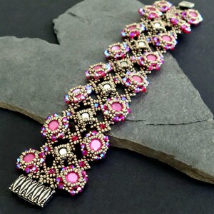 K&ouml;nnte beinhalten: Ein rosafarbenes und silbernes Armband mit einem silbernen Verschluss. Das Armband ist aus kleinen runden Perlen gefertigt und hat ein geometrisches Muster. "Beaded by Christiane Toll" steht unter dem Armband.