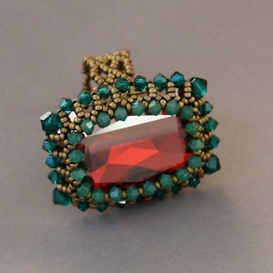 Può includere: Un anello con una grande pietra preziosa rossa circondata da un bordo di perline verdi e filo d'oro.