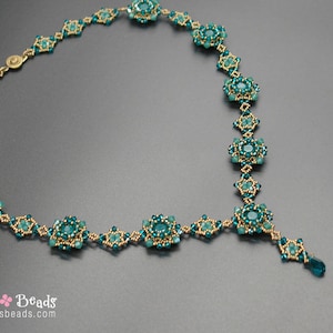 Pu&ograve; includere: Collana artigianale con perline color teal e oro, con design floreale e pendente a goccia. La chiusura &egrave; dorata. La collana &egrave; esposta su una superficie grigia. Il nome del marchio "Sidonia's Beads" &egrave; visibile.