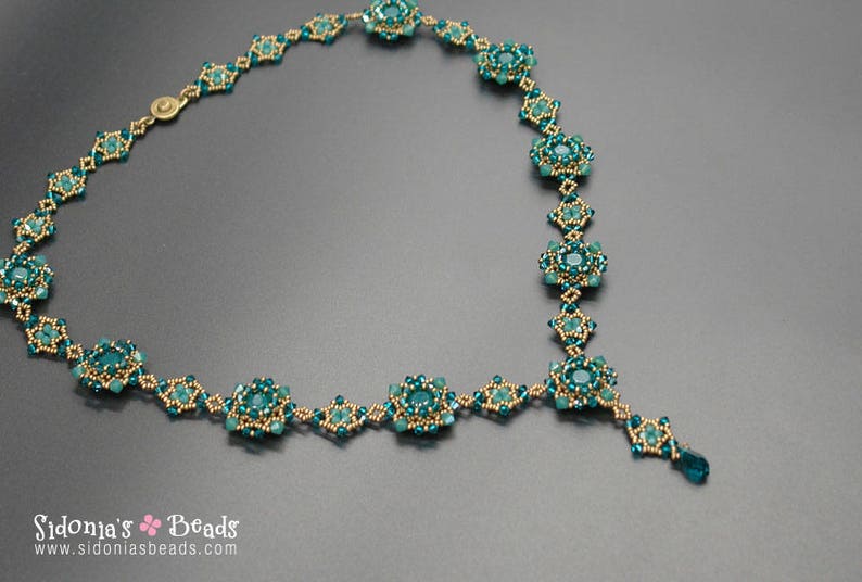 Pu&ograve; includere: Collana artigianale con perline color teal e oro, con design floreale e pendente a goccia. La chiusura &egrave; dorata. La collana &egrave; esposta su una superficie grigia. Il nome del marchio "Sidonia's Beads" &egrave; visibile.
