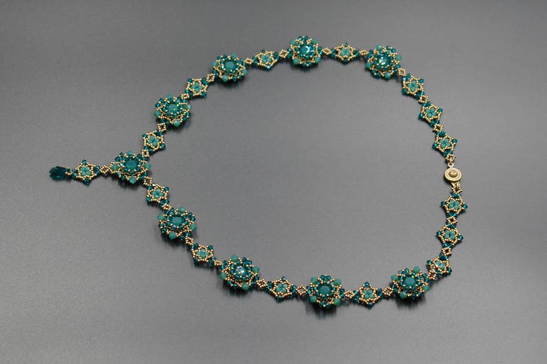 Pu&ograve; includere: Un'elegante collana con perline color teal e dettagli dorati. La collana presenta un design floreale con un pendente centrale e una delicata chiusura. L'intricato lavoro di perline crea un accessorio sofisticato e accattivante.