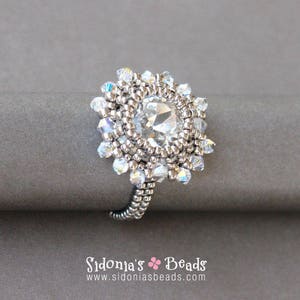 May include: A silver ring featuring a large, clear crystal at its centre, surrounded by smaller crystals. The ring is displayed against a grey backdrop.