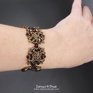 May include: A gold and burgundy beaded bracelet with a square design. The bracelet is made with small beads and has a delicate, intricate look.